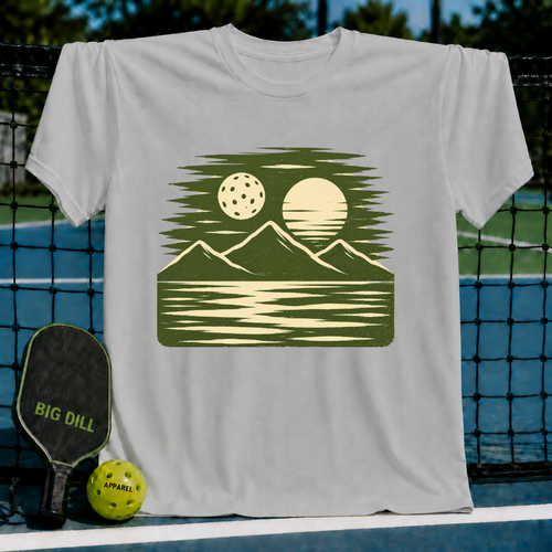 Cream unisex t-shirt featuring a forest green woodblock-style print of a pickleball, sun, mountains, and water reflection.