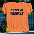 Vivid orange unisex t-shirt featuring bold navy text reading 7 FEET OF REGRET, displayed on a pickleball court net.