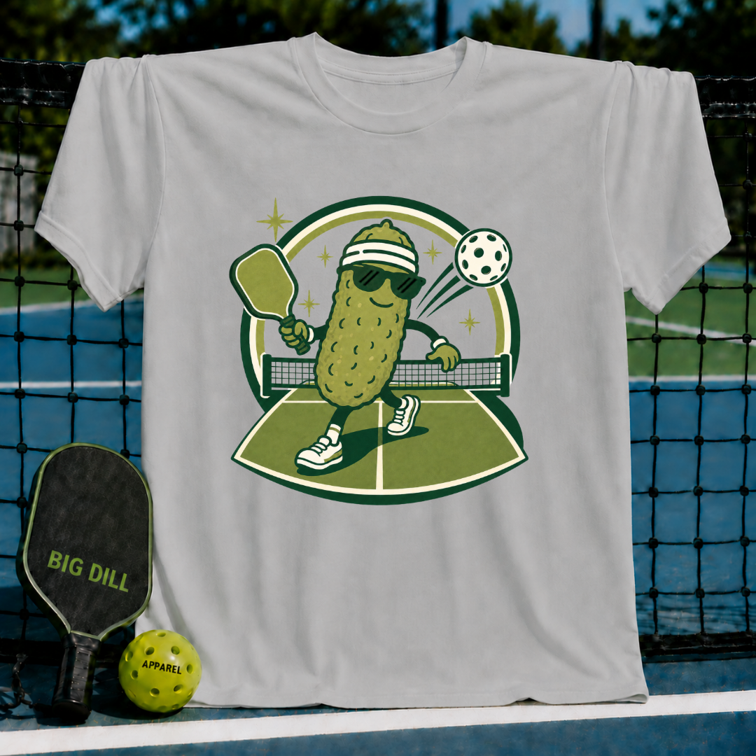 Light grey crew neck t-shirt featuring a cartoon pickle playing pickleball on a court with a paddle and ball.
