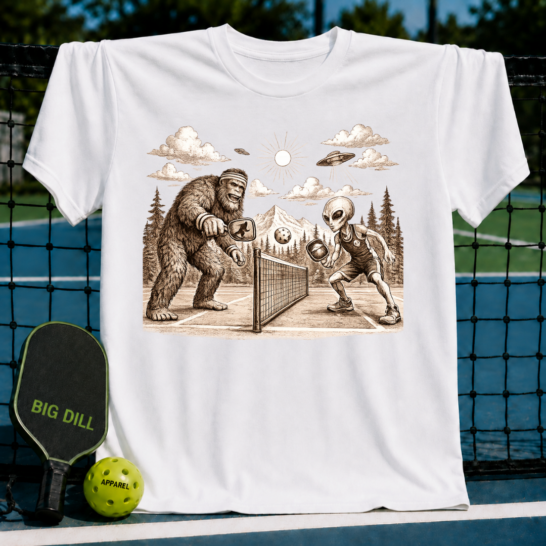 White unisex t-shirt featuring a vintage-style engraving of Bigfoot and an alien playing pickleball under a mountain sky.