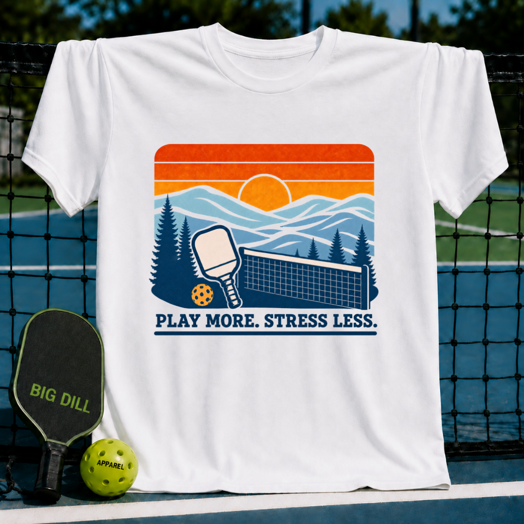 White cotton t-shirt featuring a retro sunset pickleball graphic with the text Play More. Stress Less. on a court background.