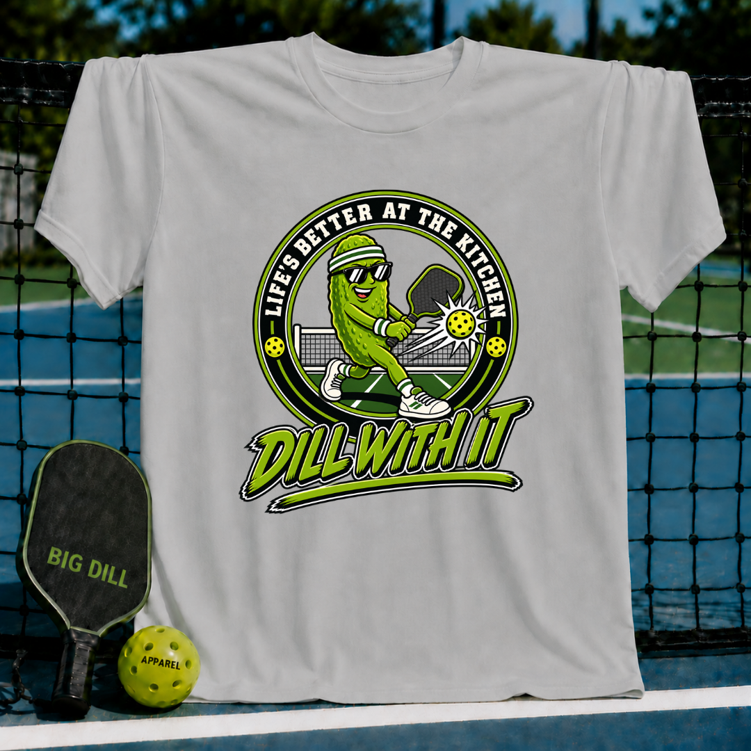 Light gray crew neck t-shirt featuring a cartoon pickle playing pickleball with the text Dill With It and Life's Better at the Kitchen.