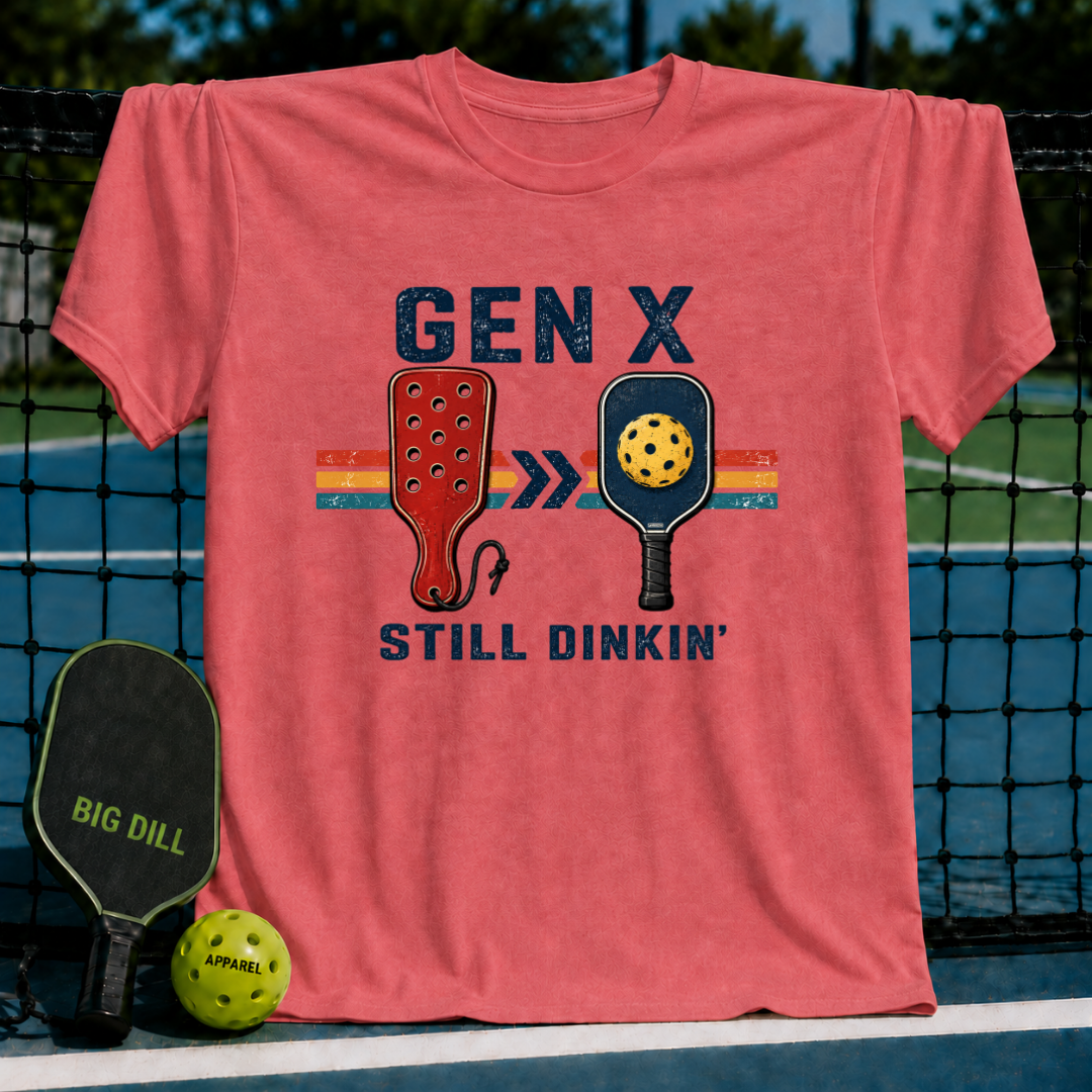 Coral pink unisex t-shirt featuring a retro Gen X Still Dinkin' graphic with vintage paddles on a pickleball court background.