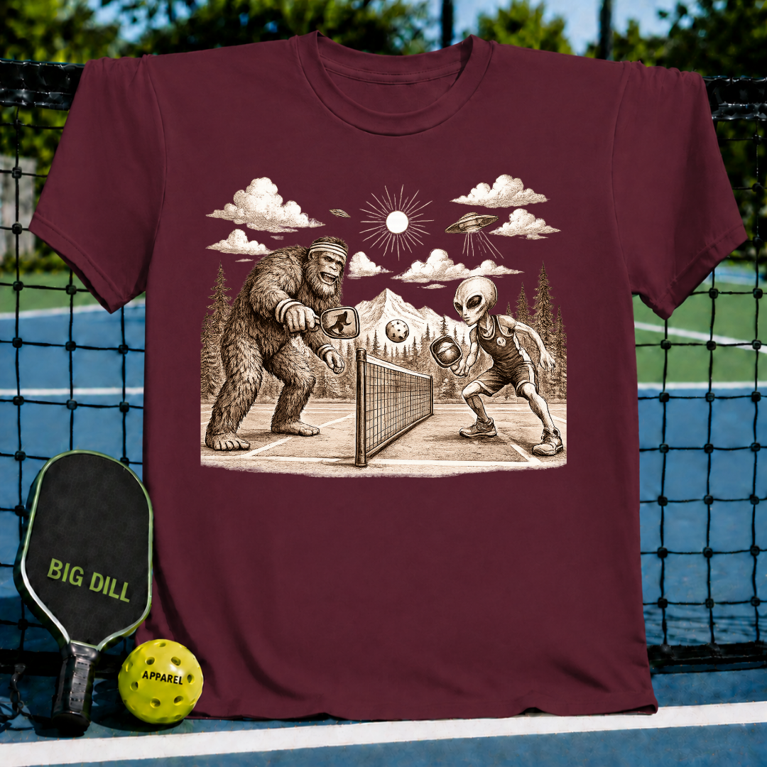 Maroon unisex t-shirt featuring a vintage-style graphic of Bigfoot and an alien playing pickleball under a mountain sky.
