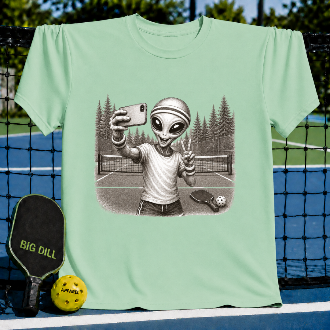 Sage green unisex t-shirt featuring a vintage-style illustration of an alien taking a selfie on a pickleball court.