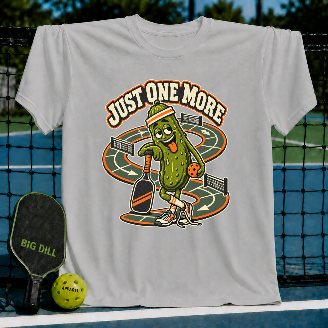 Light gray cotton t-shirt featuring a cartoon pickle playing pickleball with the text Just One More on a court background.