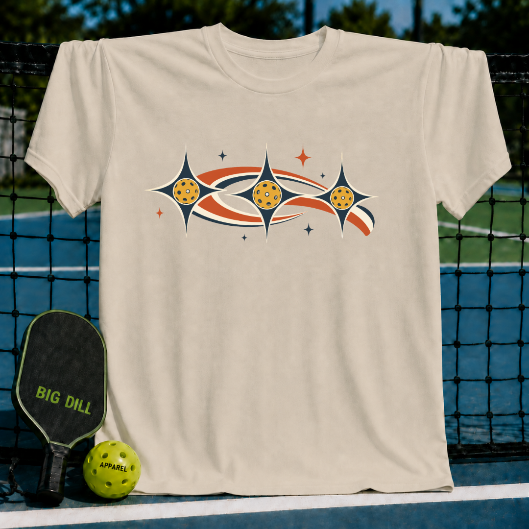 Beige unisex t-shirt featuring a retro celestial pickleball graphic with stars and orbiting earth-tone swooshes on a court.