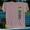 Mauve unisex T-shirt featuring a vertical green Pickleball graphic and paddle design, displayed on a pickleball court net.