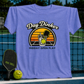 Periwinkle blue unisex t-shirt featuring a retro sunset graphic of a pickleball paddle and ball in a glass.