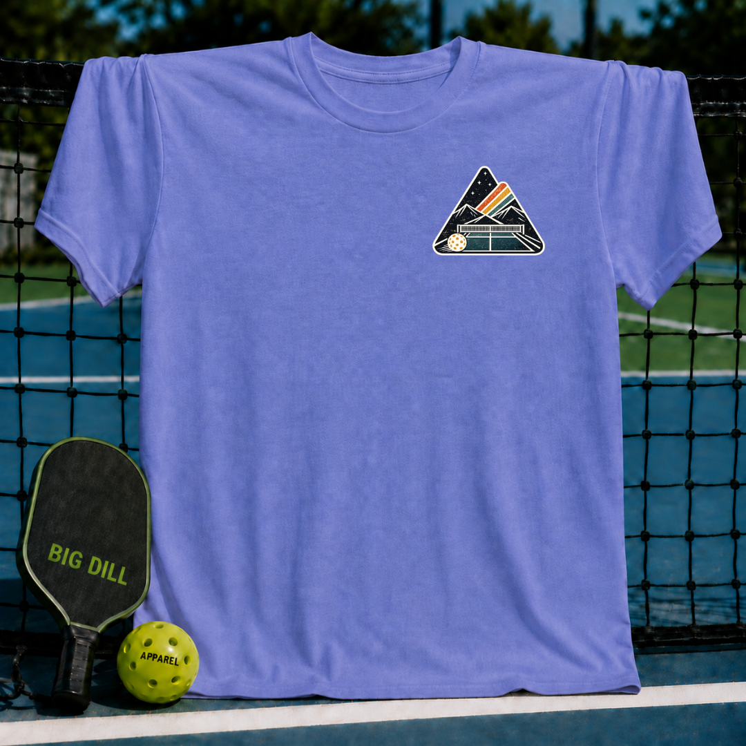 Periwinkle unisex t-shirt featuring a mountain and pickleball court crest graphic, displayed on a pickleball court net.