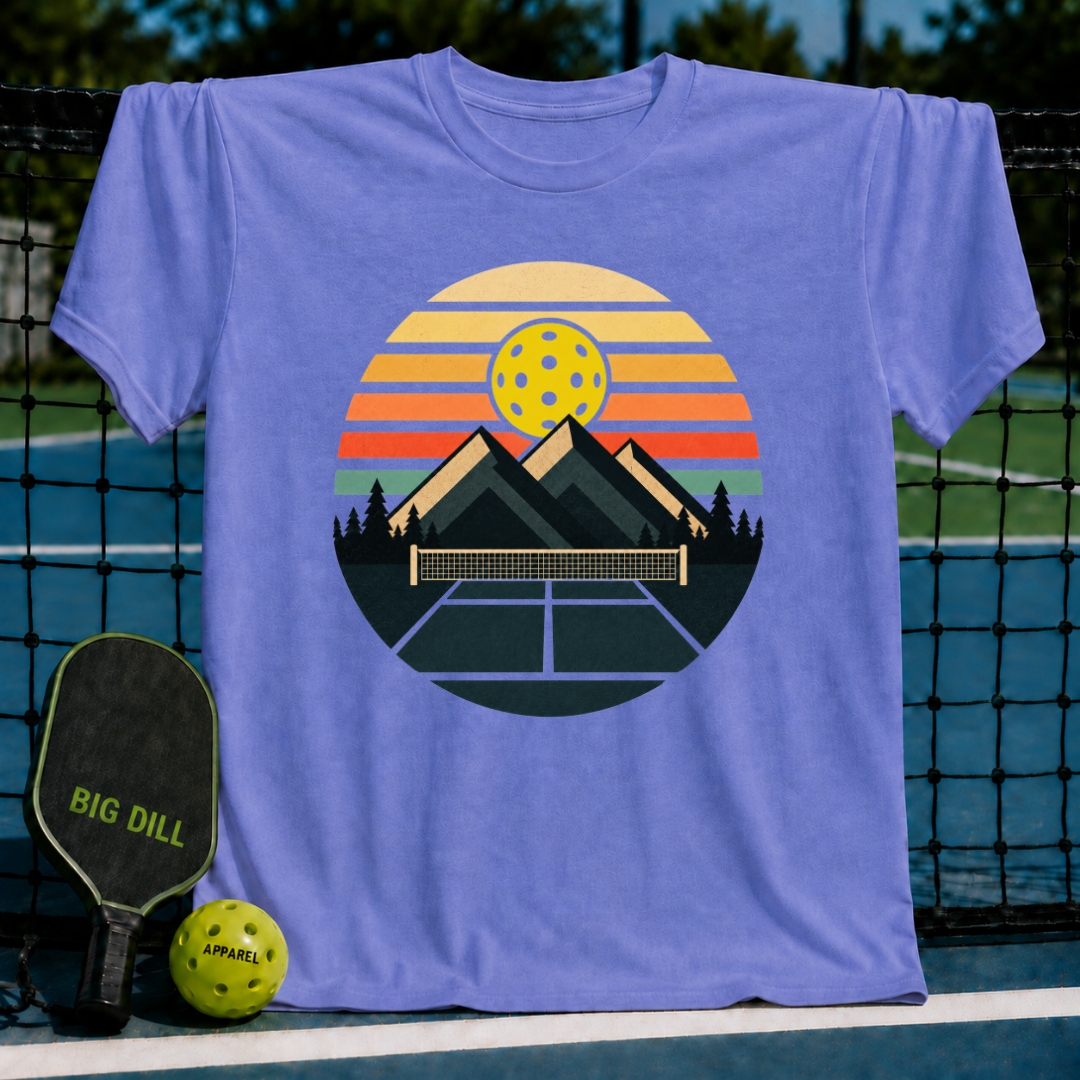 Periwinkle blue t-shirt featuring a retro circular graphic of mountains, a pickleball net, and a sunset on a pickleball court.