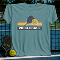 Vintage blue t-shirt featuring a retro pickleball paddle and ball graphic with striped accents on a court background.