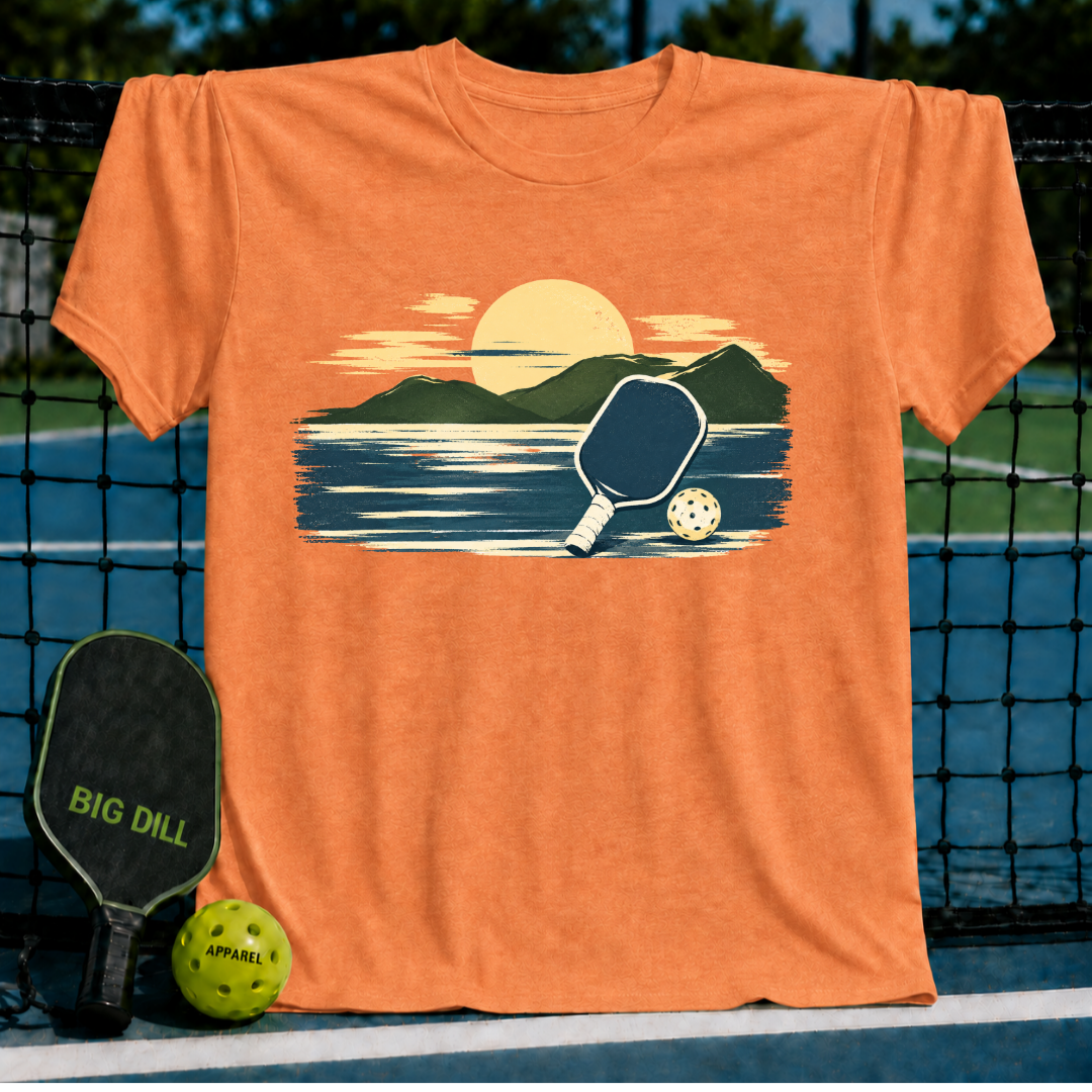 Orange unisex t-shirt featuring a painterly mountain, water, and pickleball graphic, displayed on a court net.