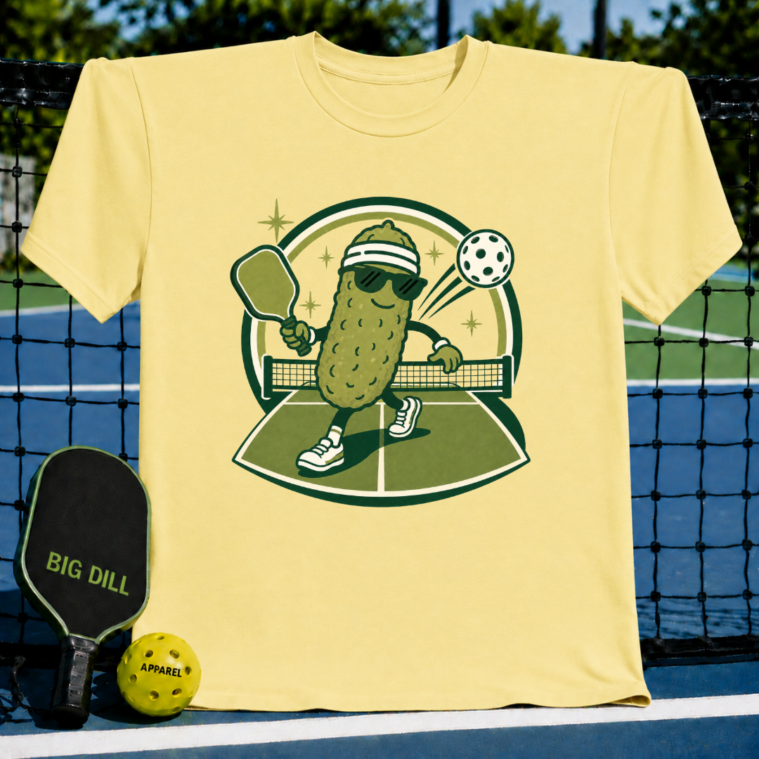 Light yellow crewneck t-shirt featuring a cartoon pickle playing pickleball with sunglasses and a headband.