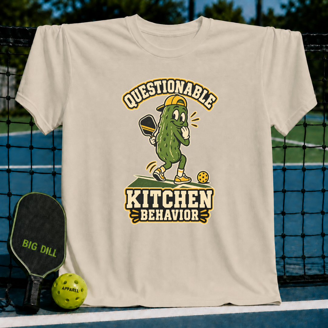 Beige crewneck t-shirt featuring a cartoon pickle playing pickleball with the text Questionable Kitchen Behavior.