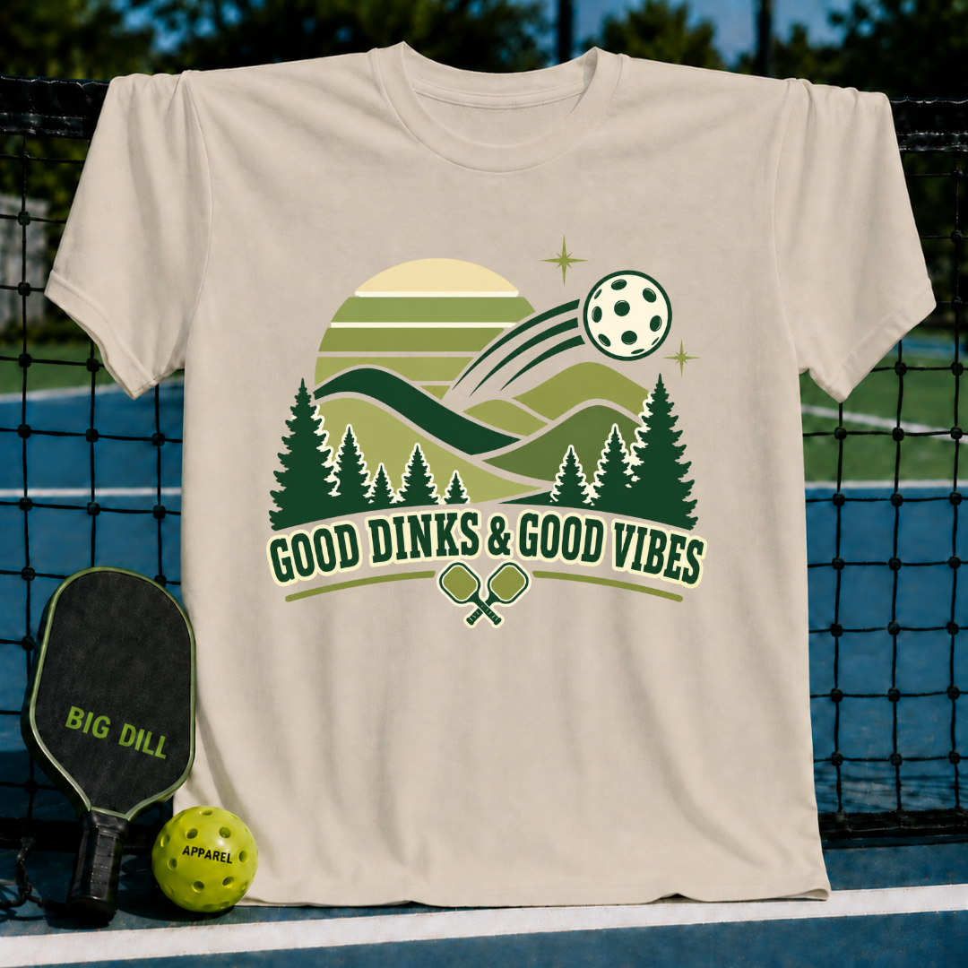 Beige short-sleeve t-shirt featuring a green pickleball-themed landscape graphic with the text Good Dinks & Good Vibes.