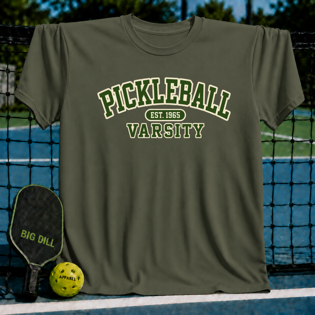 Olive green crewneck t-shirt featuring white varsity-style Pickleball Est. 1965 text, displayed on a pickleball court net.