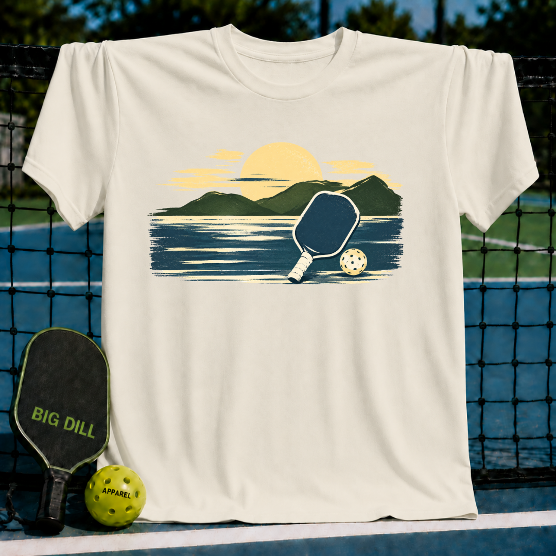 Cream-colored unisex t-shirt featuring a painterly mountain and pickleball graphic in navy and gold tones.