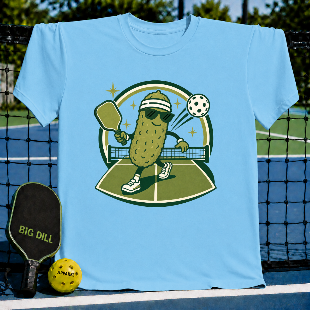 Light blue crew neck t-shirt featuring a cartoon pickle wearing sunglasses and a headband playing pickleball.