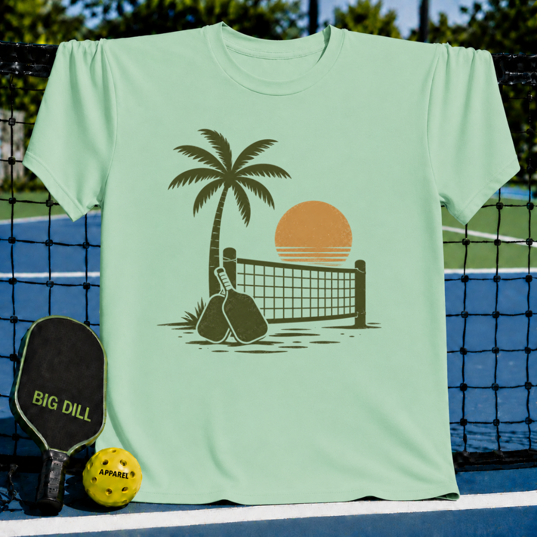 Light green unisex t-shirt featuring a brown palm tree, pickleball net, and paddle graphic against a sunset background.