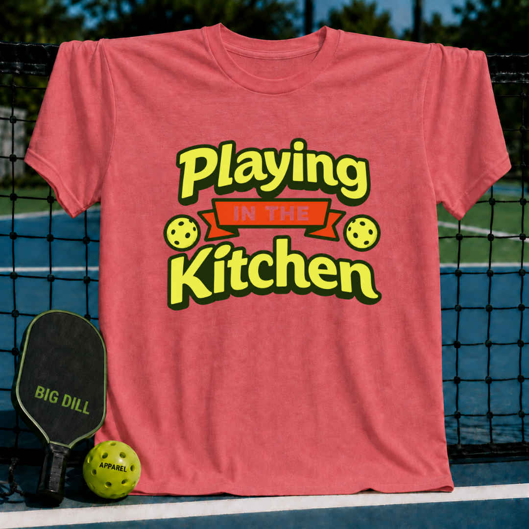 Coral pink crewneck t-shirt featuring Playing in the Kitchen text with pickleball graphics, displayed on a pickleball court.