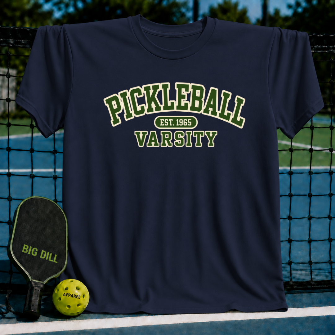 Navy blue varsity-style t-shirt with green and cream Pickleball text, displayed on a court with a paddle and ball.