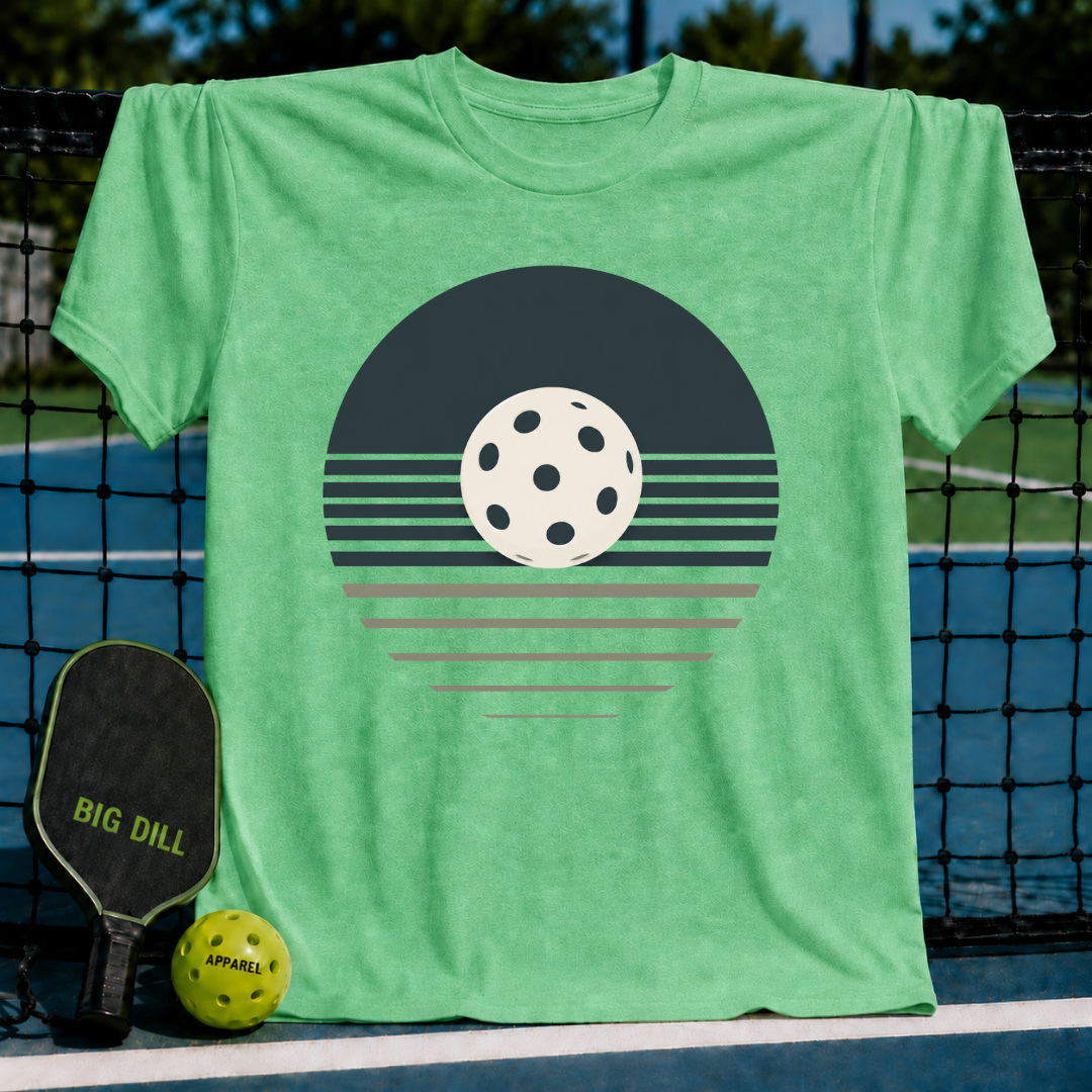 Soft green unisex t-shirt featuring a retro pickleball sunset graphic in charcoal and cream, displayed on a court net.