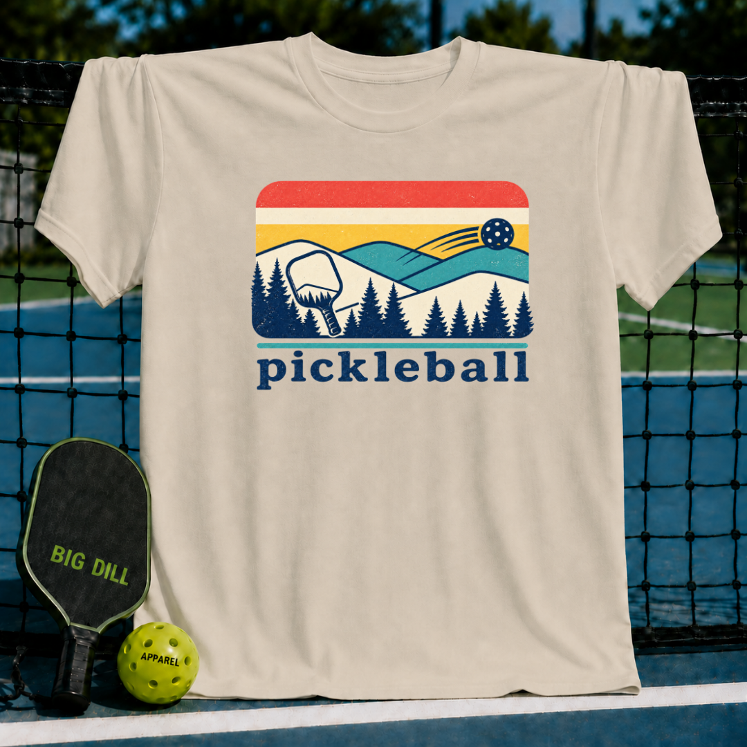 Cream unisex t-shirt featuring a retro mountain, pine forest, and pickleball graphic with red, yellow, and blue accents.