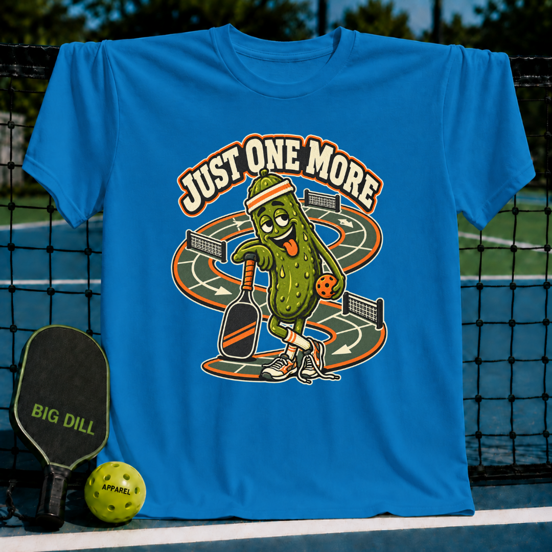Bright blue crewneck t-shirt featuring a cartoon pickle character playing pickleball with the text Just One More.