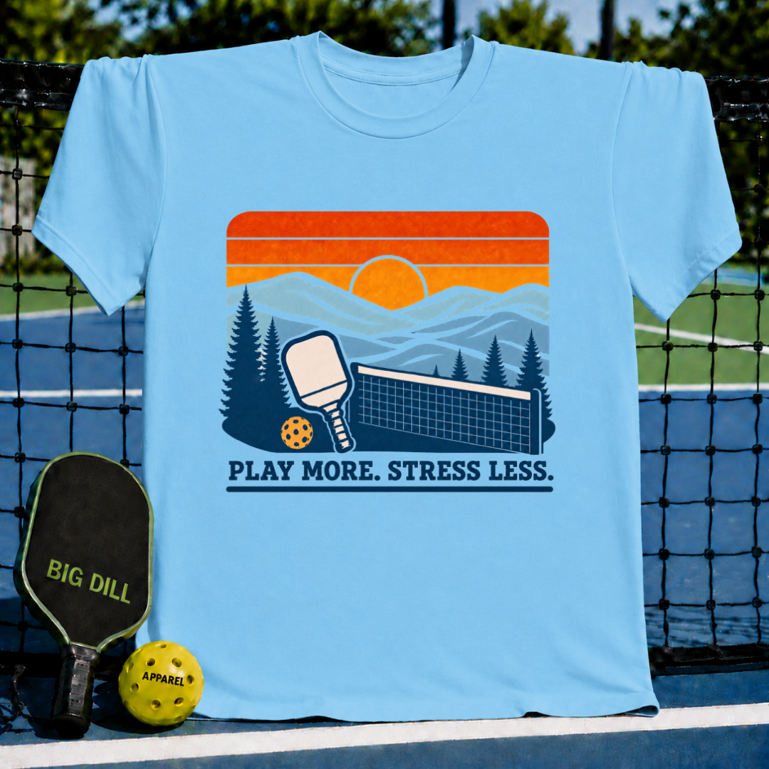 Light blue crewneck t-shirt featuring a retro sunset pickleball graphic with a paddle, ball, and the text Play More. Stress Less.