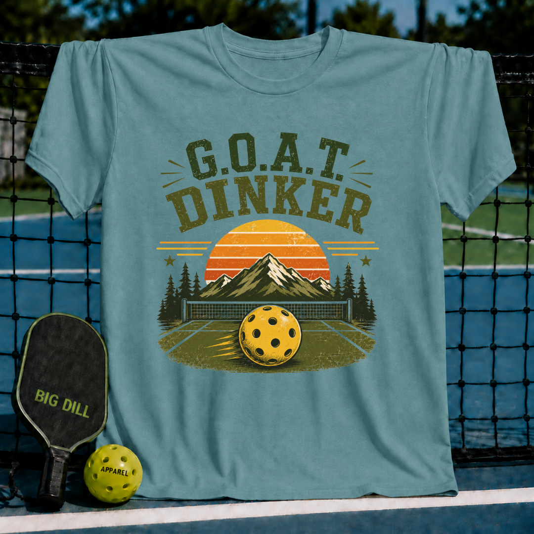 Periwinkle blue unisex t-shirt featuring a retro pickleball graphic with a sunset, mountains, and G.O.A.T. DINKER text.