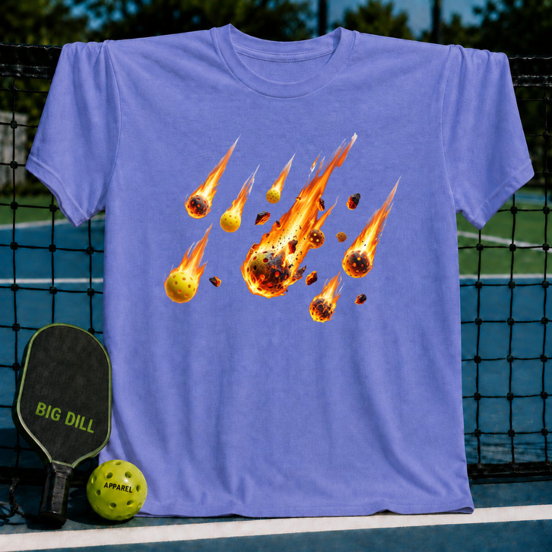 Periwinkle blue unisex t-shirt featuring a flaming pickleball meteor shower graphic on the chest, displayed on a pickleball court.
