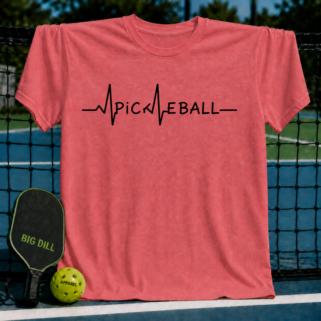 Coral unisex t-shirt featuring a black EKG heartbeat graphic that spells out PICKLEBALL, displayed on a pickleball court.