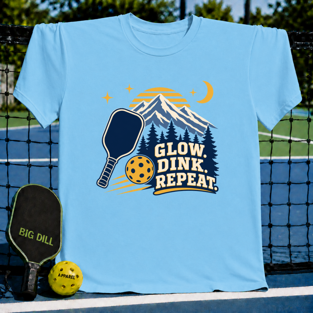 Light blue t-shirt featuring a mountain, pickleball paddle, and ball graphic with the text Glow, Dink, Repeat.