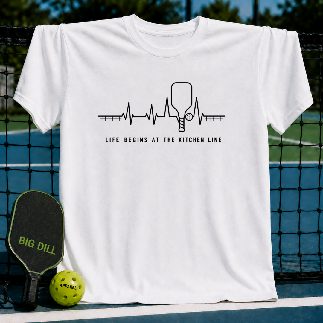 White unisex t-shirt featuring a black heartbeat graphic with a pickleball paddle and text Life Begins At The Kitchen Line.