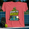 Coral pink t-shirt featuring a cartoon pickle character playing pickleball with the text Down the Middle Solves the Riddle.
