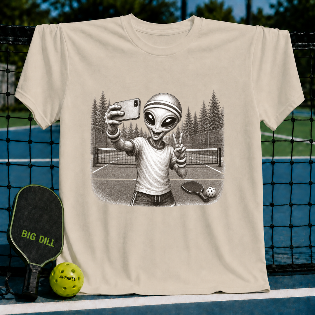 Light beige unisex t-shirt featuring a vintage-style illustration of an alien taking a selfie on a pickleball court.