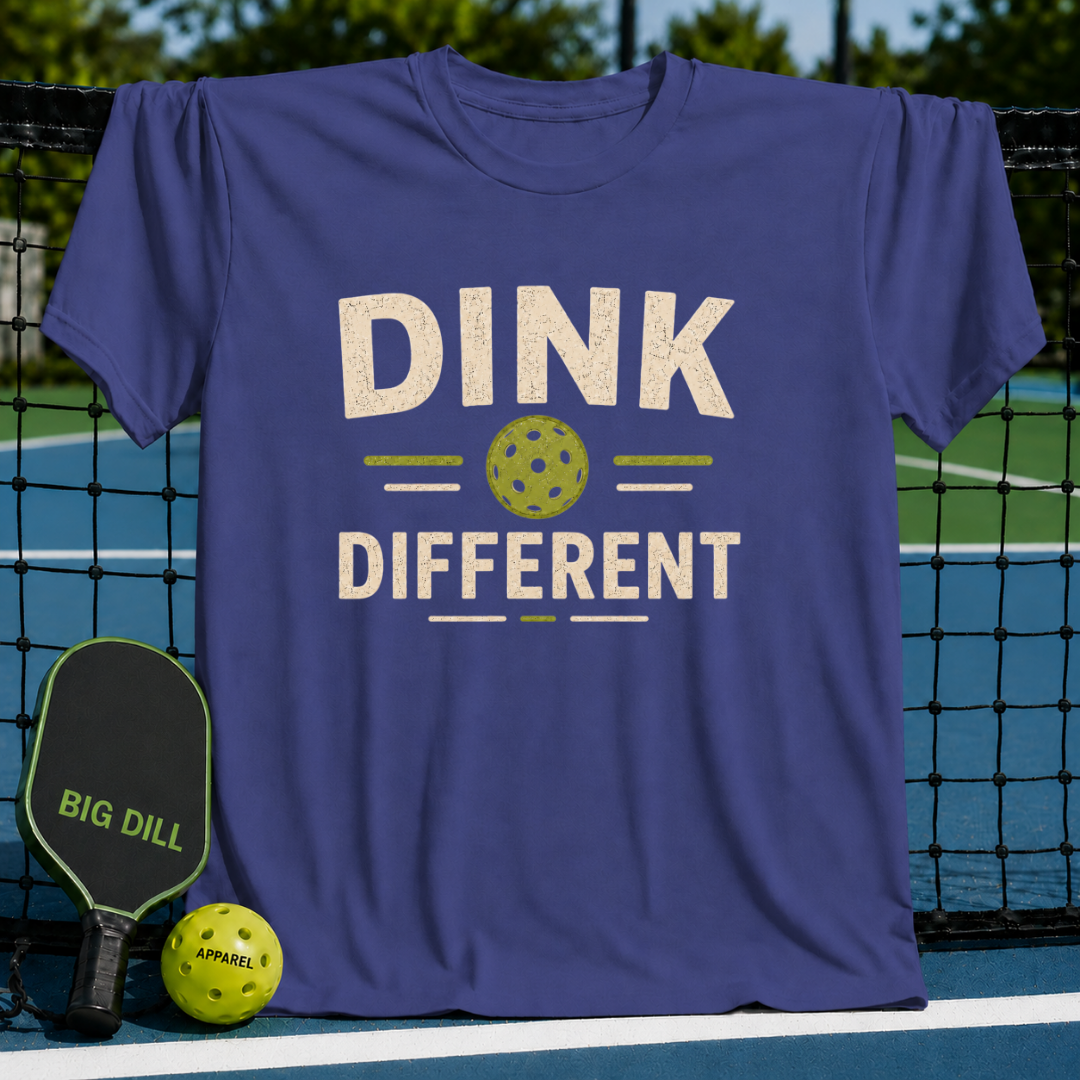 Blue Dink Different pickleball t-shirt with a distressed cream and green graphic, displayed on a pickleball court net.
