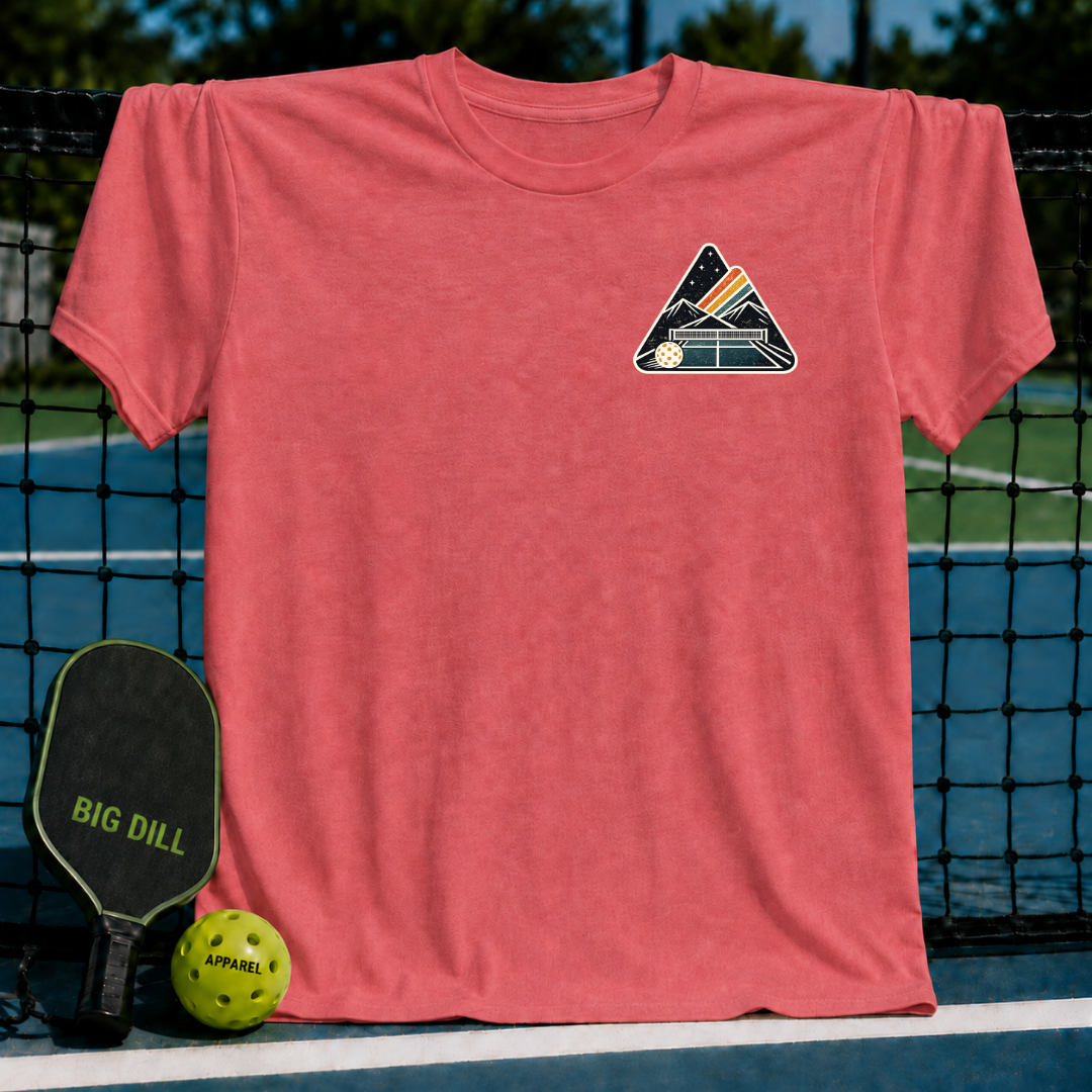 Coral unisex t-shirt featuring a mountain and pickleball court crest graphic, displayed on a pickleball court net.