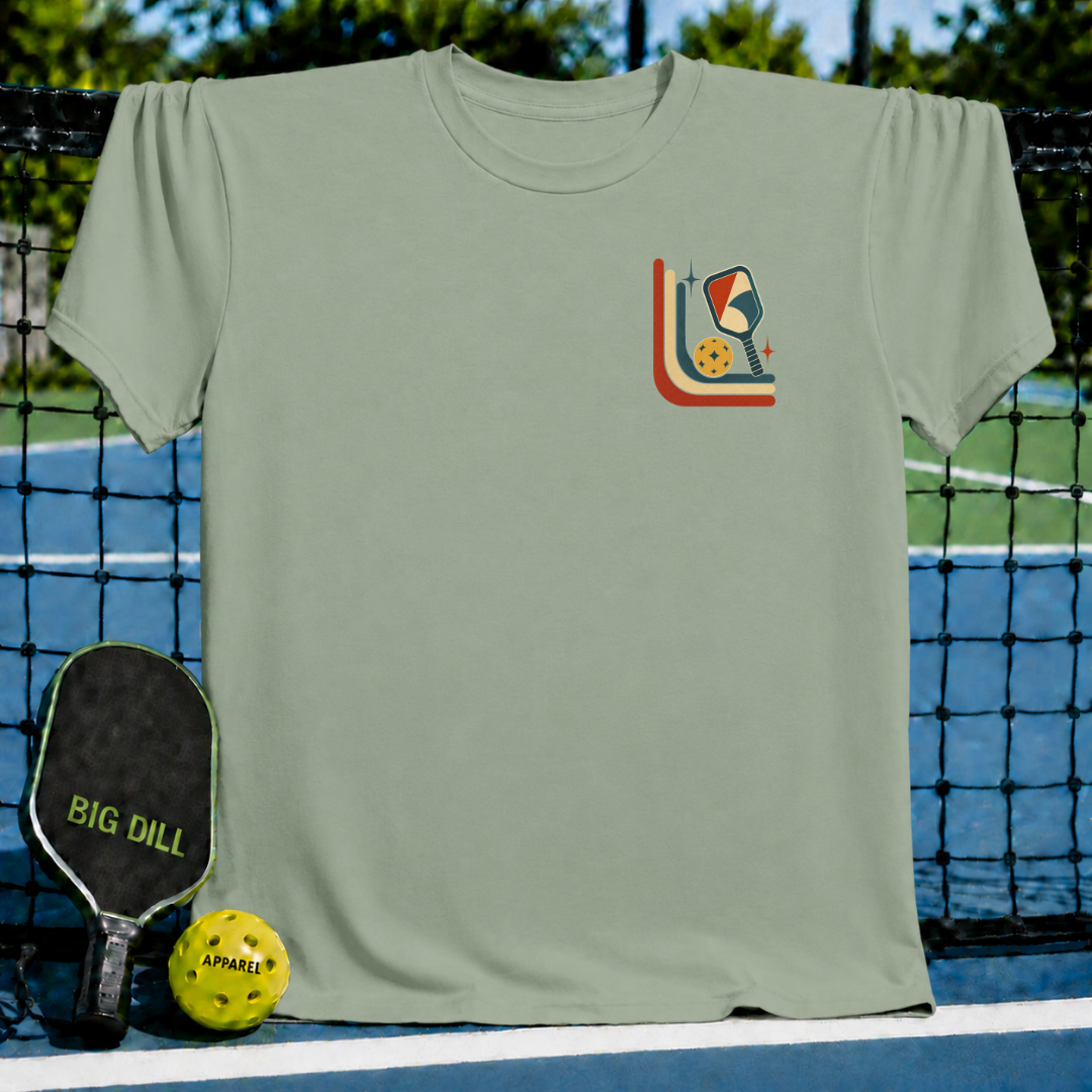 Sage green unisex t-shirt featuring a retro mid-century pickleball graphic on the chest, displayed on a pickleball court.