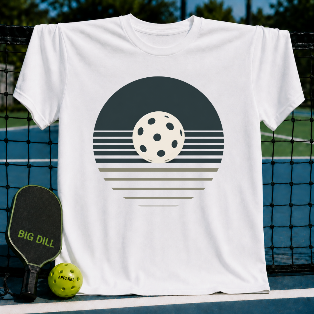 White unisex t-shirt featuring a retro charcoal and cream pickleball sunset graphic, displayed on a pickleball court.