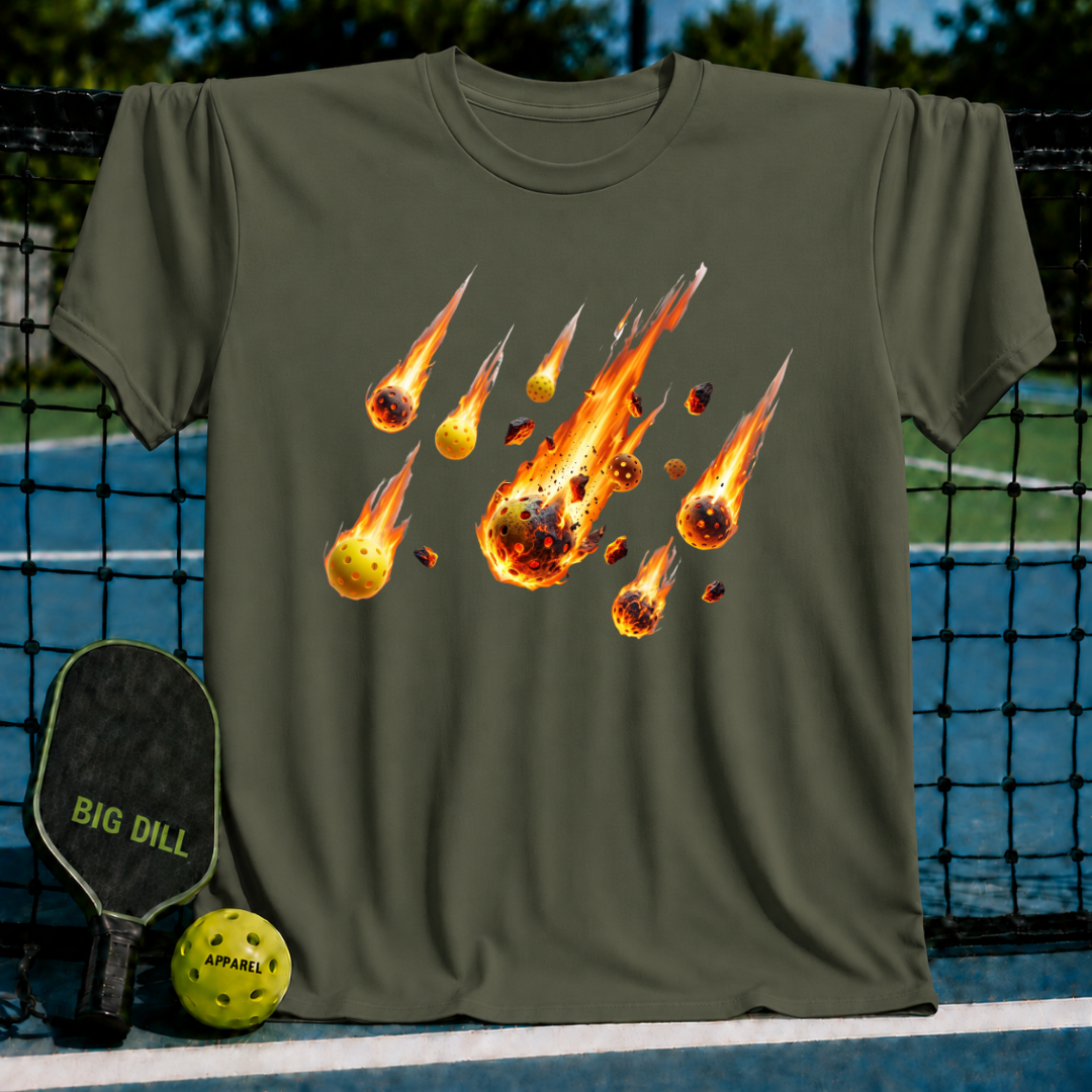 Olive green unisex t-shirt featuring a flaming pickleball meteor shower graphic on the chest, displayed on a pickleball court.