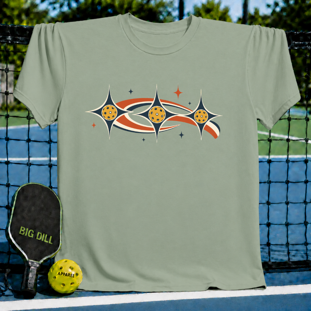 Sage green unisex t-shirt featuring a retro celestial pickleball graphic with stars and orbiting earth-tone shapes.