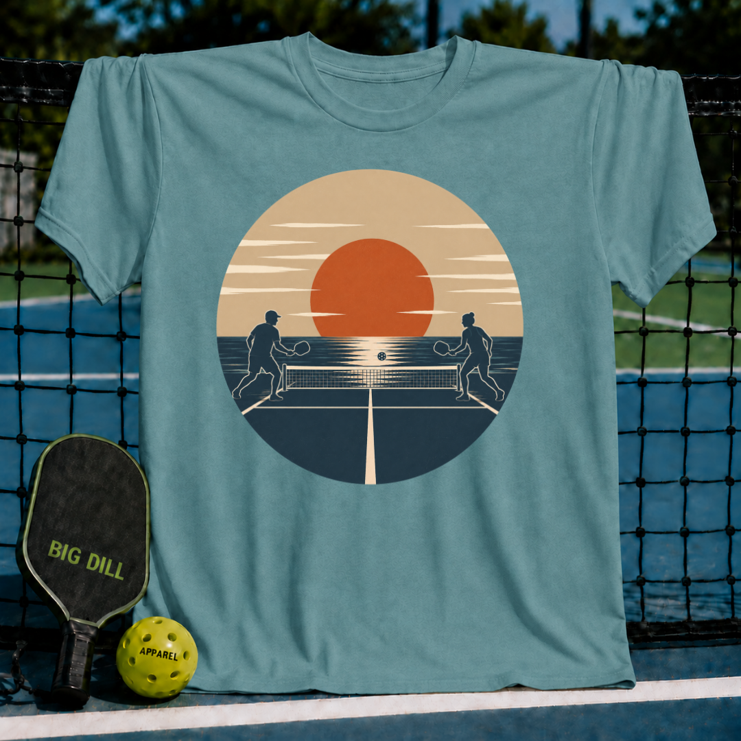 Blue-green unisex t-shirt featuring a retro sunset pickleball court silhouette graphic, displayed on a court net.