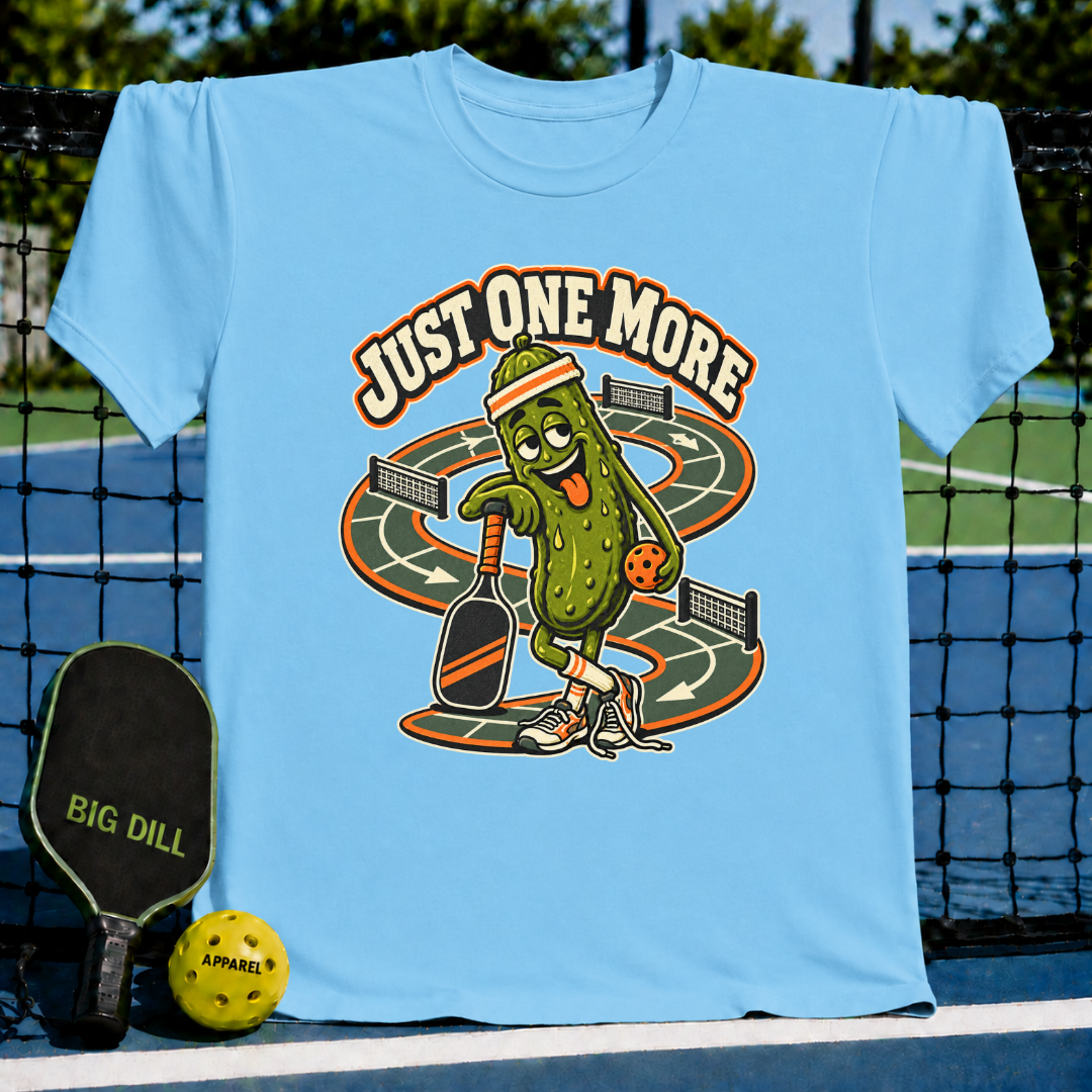 Light blue crew neck t-shirt featuring a cartoon pickle character playing pickleball with the text Just One More.