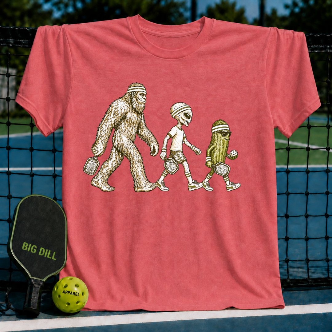 Coral red t-shirt featuring a graphic of Bigfoot, an alien, and a pickle playing pickleball on a court background.