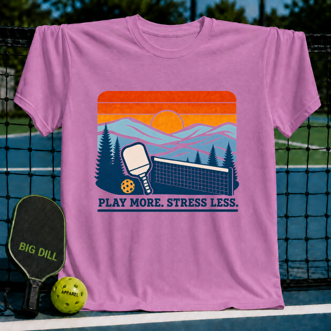 Pink cotton t-shirt featuring a retro sunset, pickleball paddle, and net graphic with the text Play More. Stress Less.