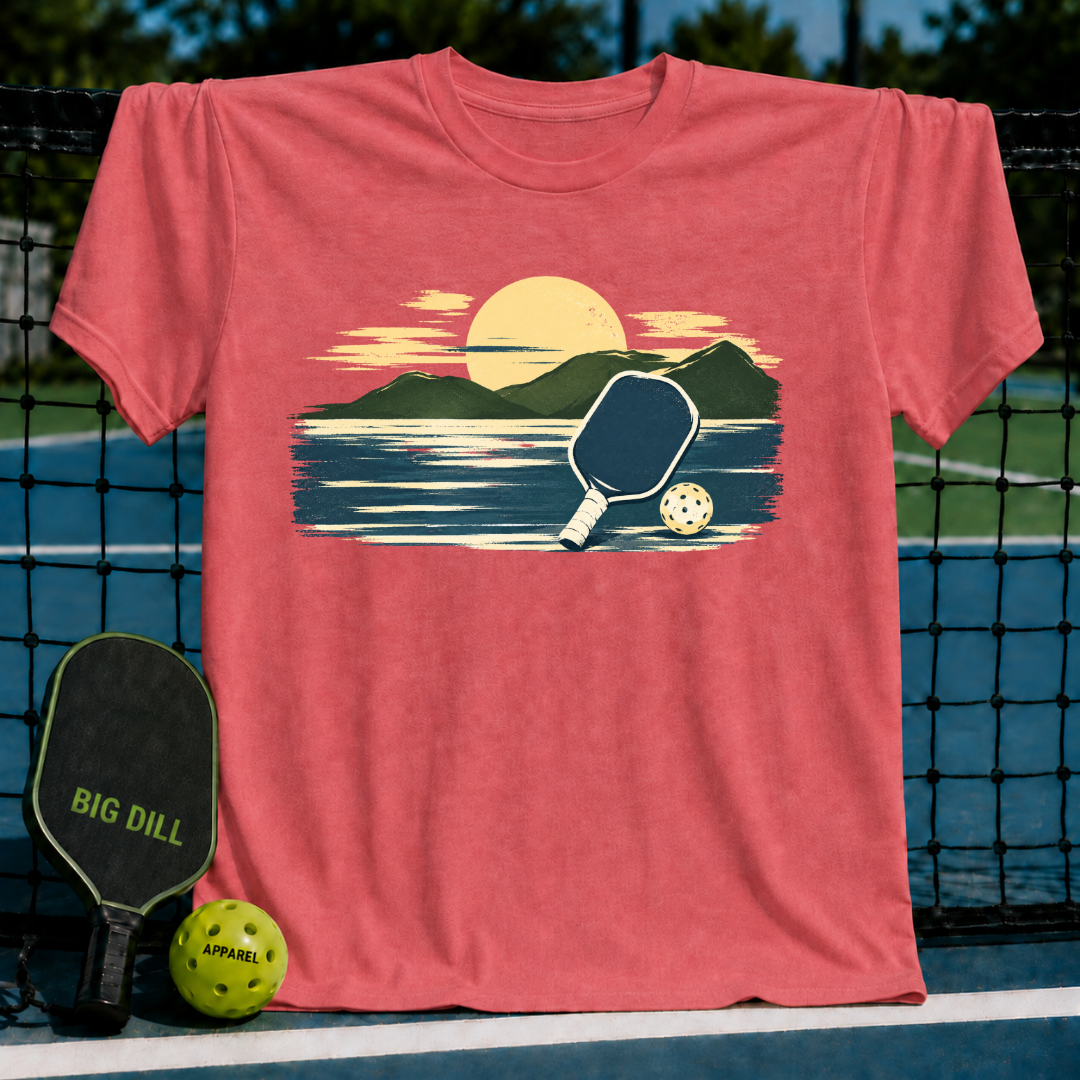 Coral-colored unisex t-shirt featuring a painterly mountain, water, and pickleball graphic against a court background.