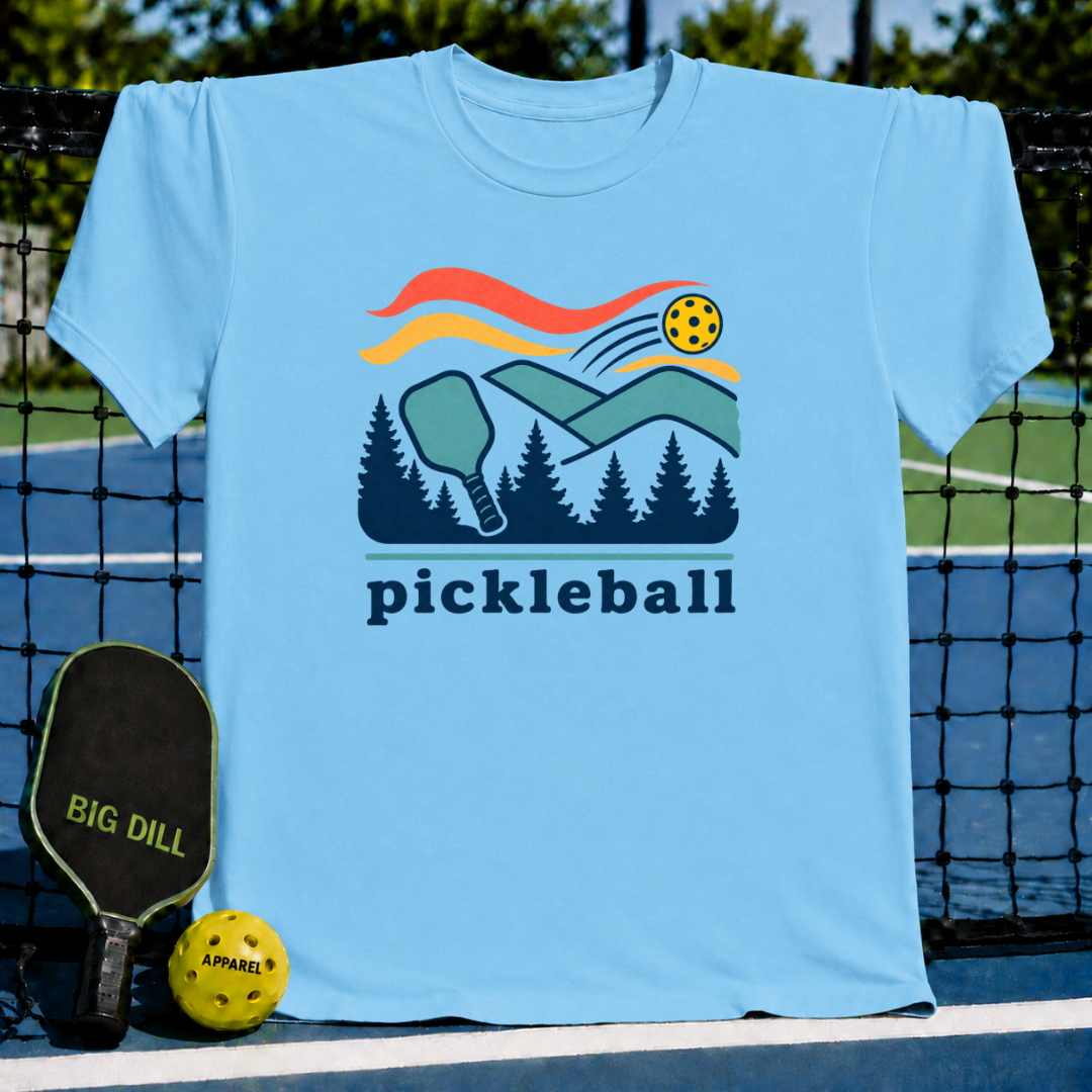 Light blue crew neck t-shirt featuring a retro pickleball, paddle, and forest landscape graphic on a pickleball court.