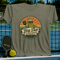 Military olive green unisex t-shirt featuring a retro pickleball mascot graphic with a pickle wearing sunglasses and a visor.